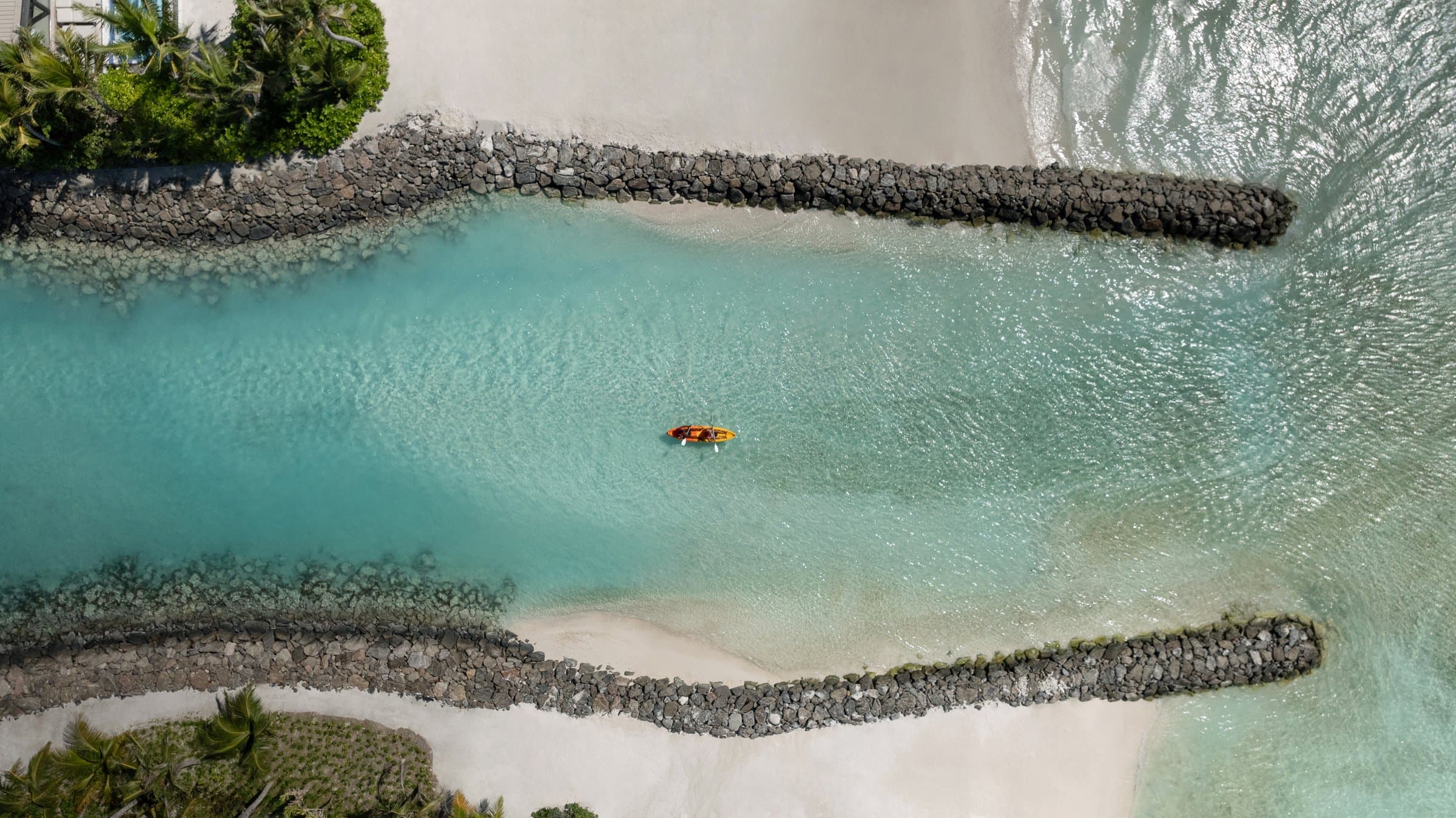 Aerial view of a channel between two stone breakwaters at SO/ Maldives, with a kayak crossing the lagoon